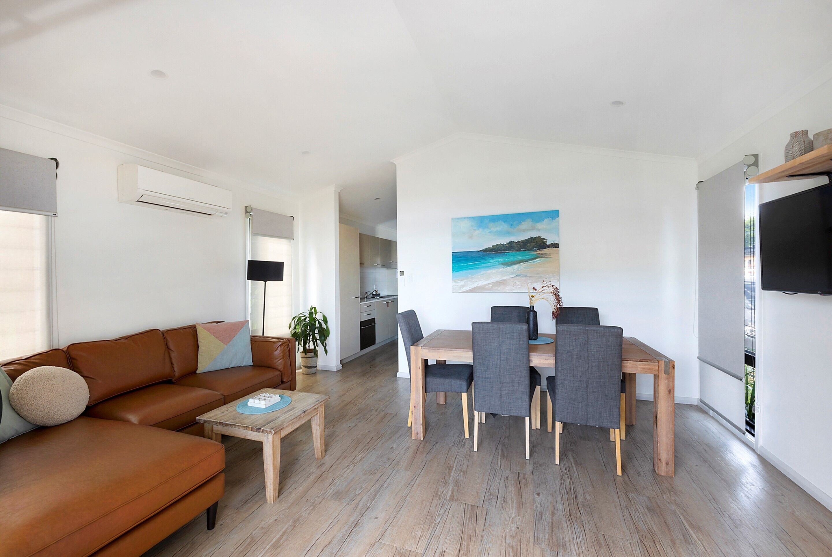 A modern living room with a brown leather sofa, a wooden coffee table, and a dining area with a wooden table and grey chairs.