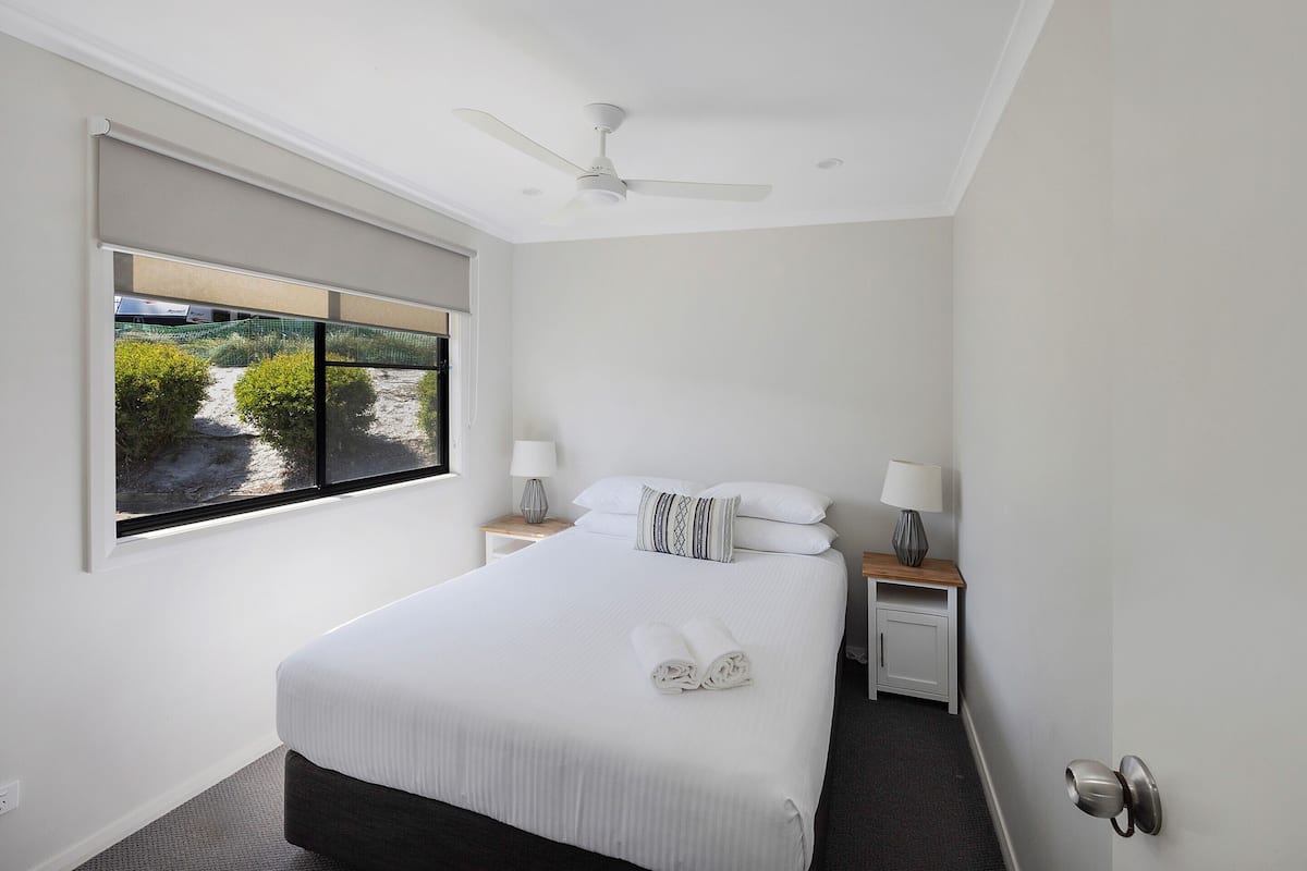 Habitación de hotel ordenada con una cama grande, ventilador de techo, ventana con vistas a la vegetación y una mesita de noche con lámpara.