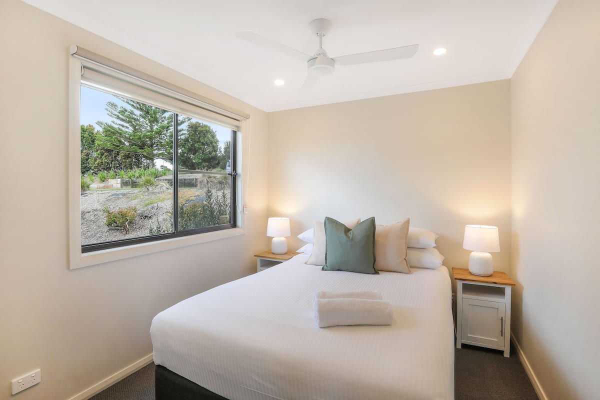 Una cama bien hecha con almohadas, una lámpara de noche y una ventana con vistas al verdor.