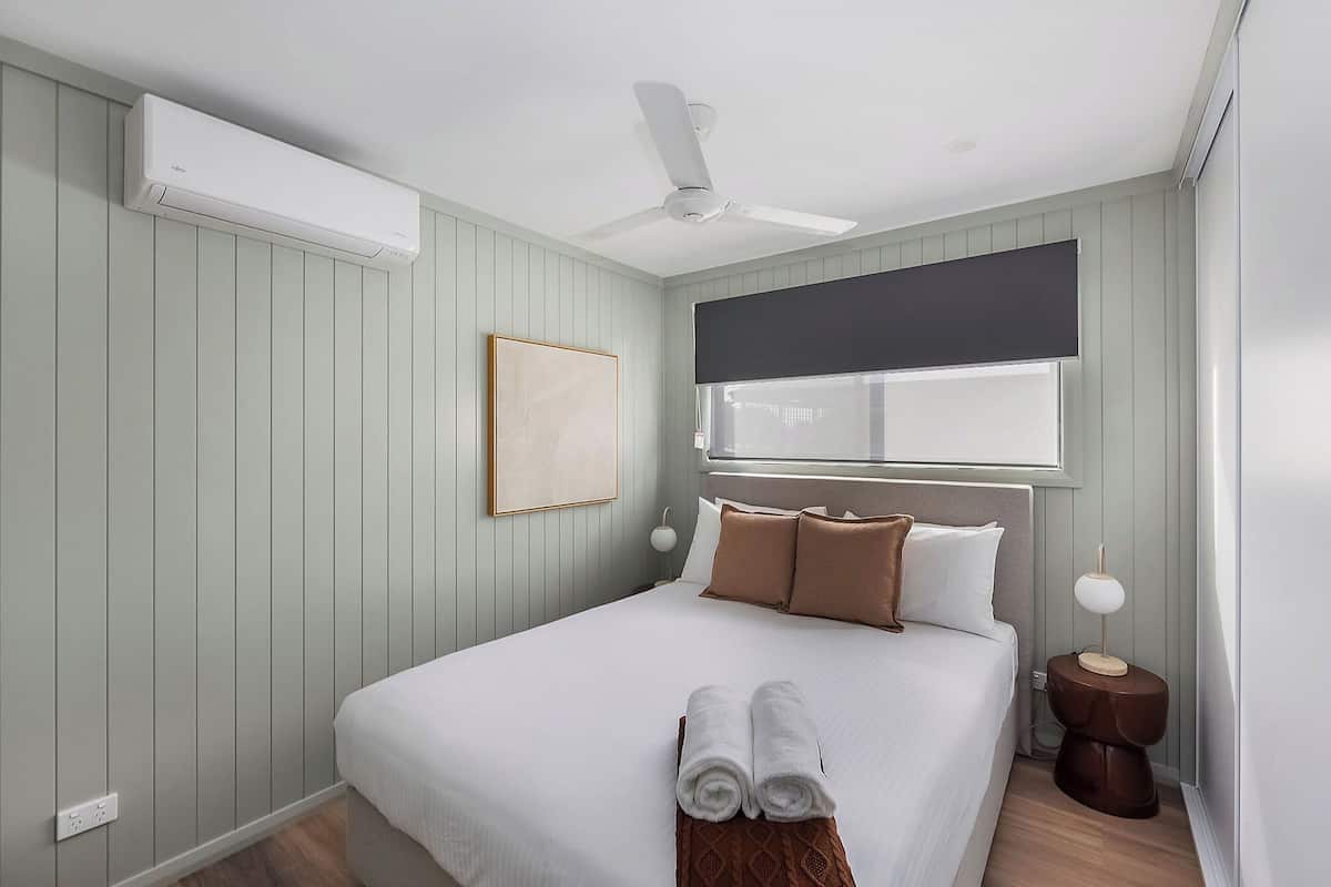 Una cama bien hecha con sábanas blancas y almohadas marrones, un ventilador de techo y una mesita de noche con una lámpara.