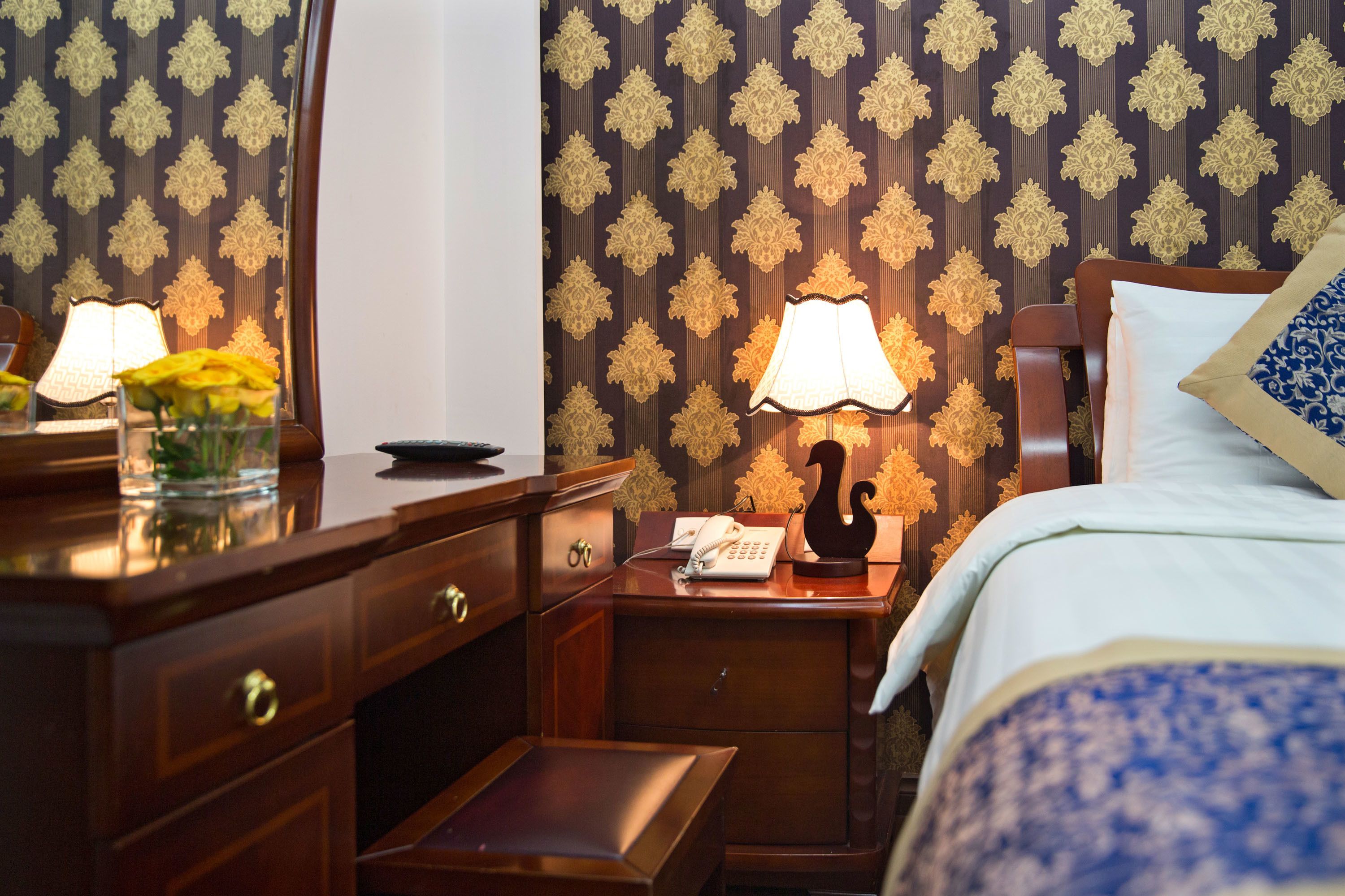 Ein Hotelzimmer mit einem Holztisch, einem Telefon, einer Lampe und einem Bett mit blau-weißer Bettdecke.