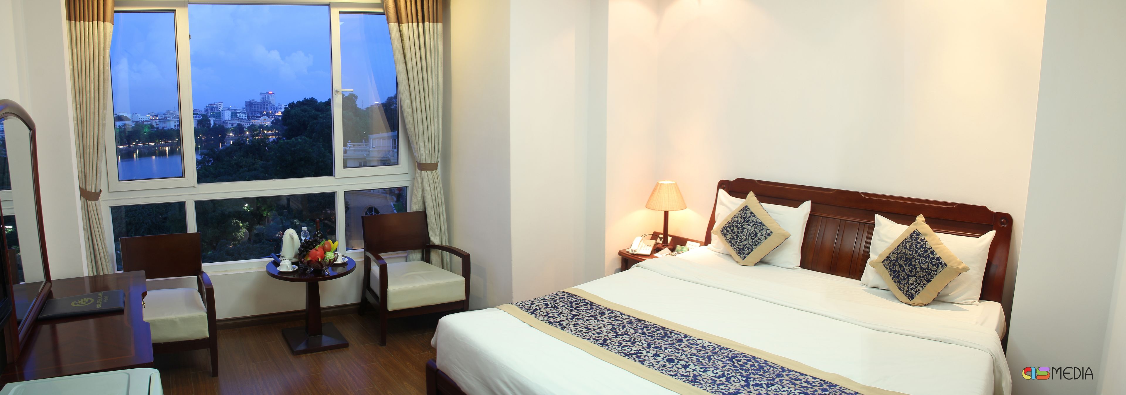 A hotel room with a bed, bedside table, lamp, chairs, and a view of a cityscape through large windows.
