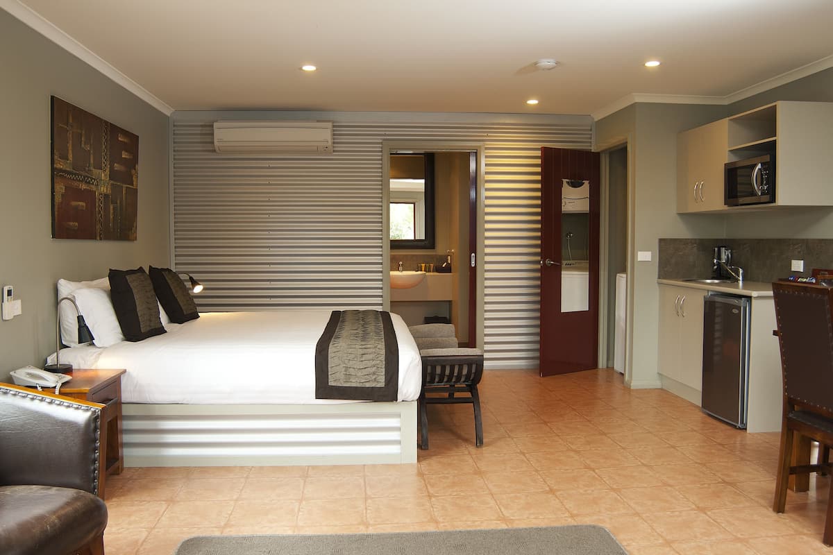 Une chambre d’hôtel moderne dotée d’un grand lit, d’un bureau, d’une chaise, d’une petite kitchenette et d’une salle de bain visible à travers une porte ouverte.