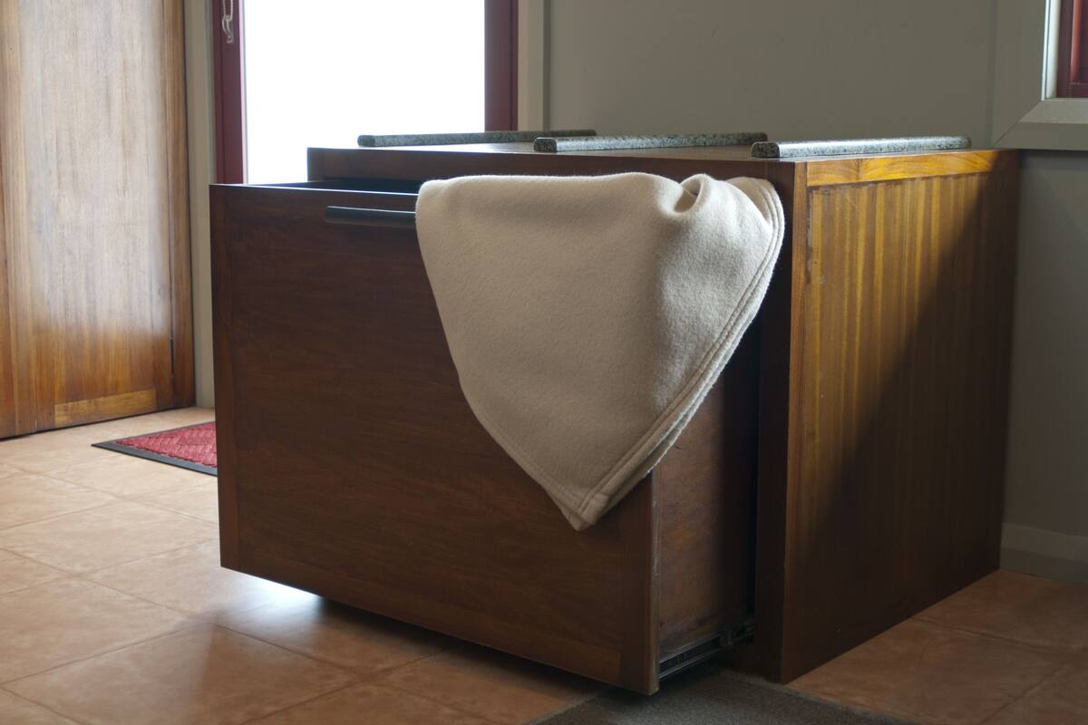Une commode en bois, sur laquelle est posée une serviette beige, se trouve dans une pièce dont le sol est carrelé et où l’on aperçoit une fenêtre au fond.