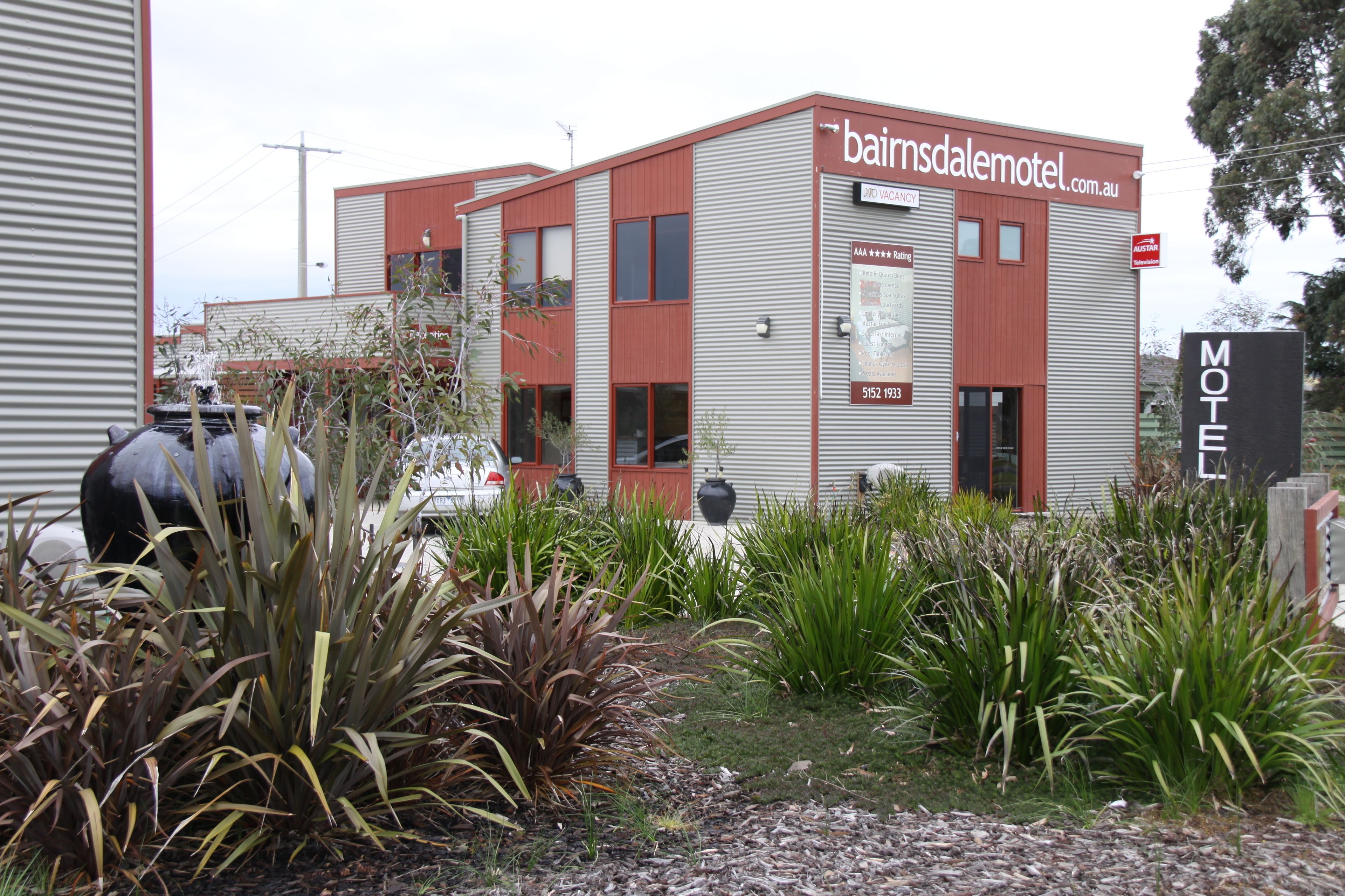Um motel com design moderno, apresentando uma placa chamativa 'bainsdalmotel.com.au', uma ampla área externa com churrasqueira e um jardim bem cuidado com diversas plantas.