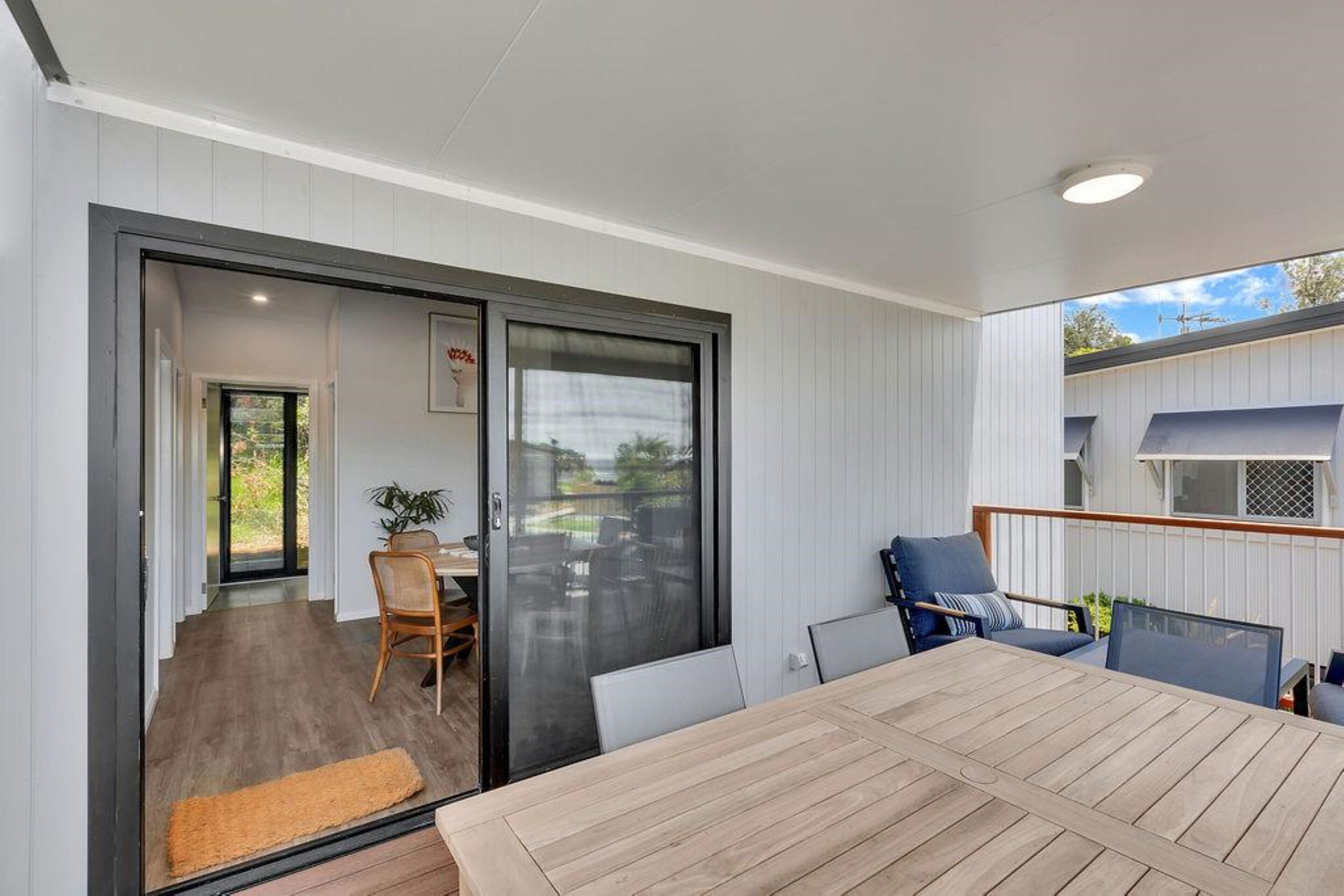 A modern outdoor dining area with a wooden table and chairs, a sliding glass door leading to a patio, and a view of a residential building.
