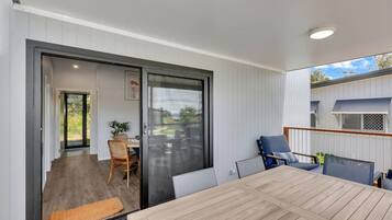 A modern outdoor dining area with a wooden table and chairs, a sliding glass door leading to a patio, and a view of a residential building.