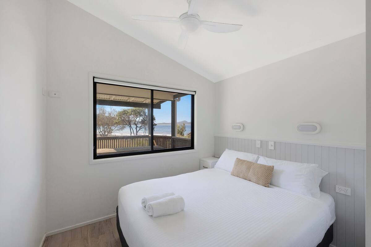 A bedroom with a large bed, a ceiling fan, a window with a view of the outdoors, and a nightstand with a lamp.