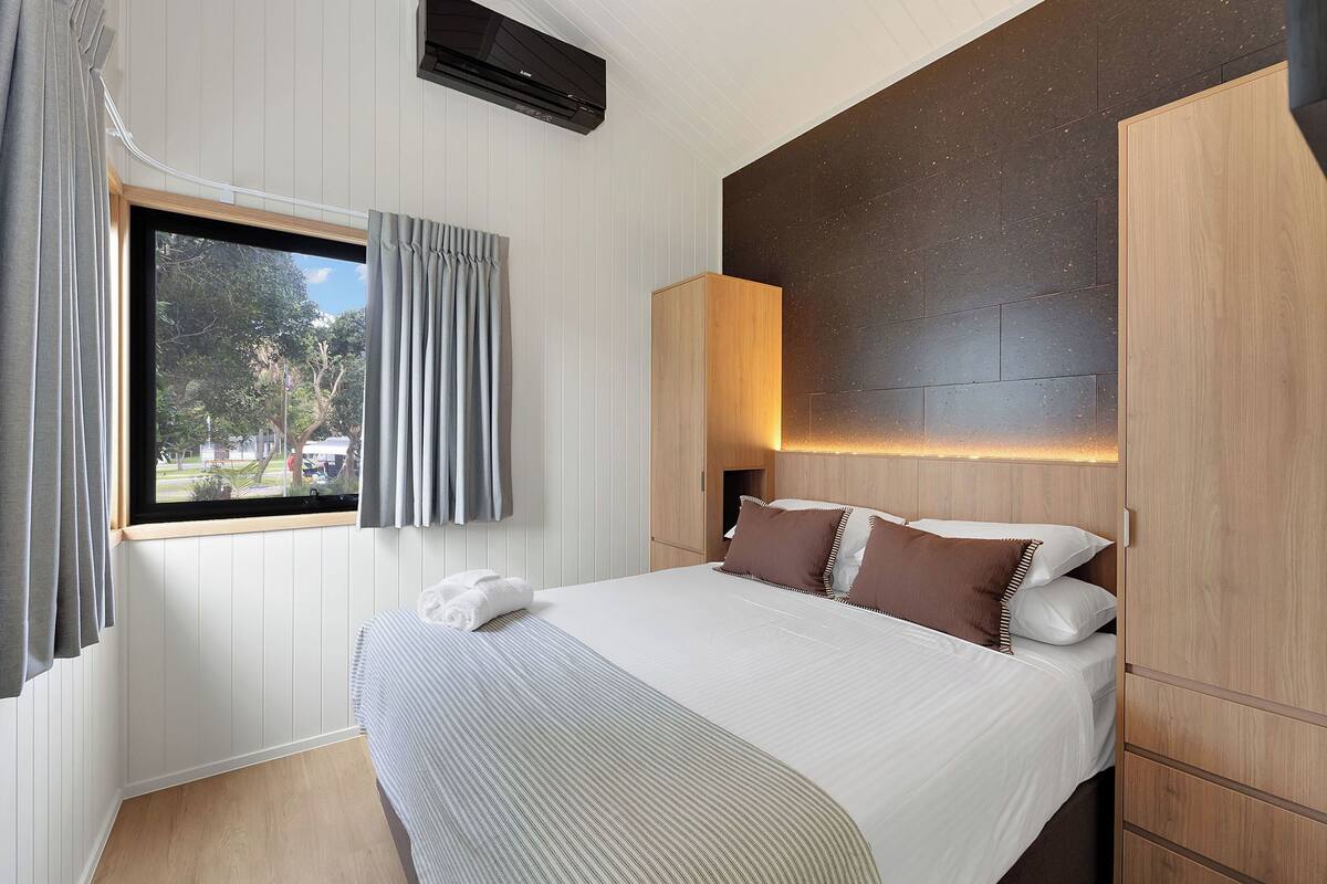 A hotel room with a bed, a window with curtains, a wall-mounted air conditioner, and a wooden headboard.