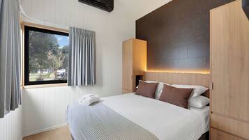 A hotel room with a bed, a window with curtains, a wall-mounted air conditioner, and a wooden headboard.