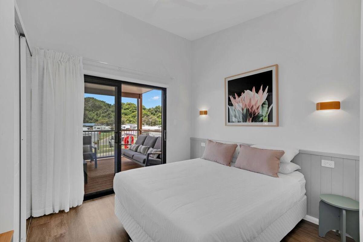 A bedroom with a large bed, a sliding glass door to a balcony, a framed picture on the wall, and a small table.