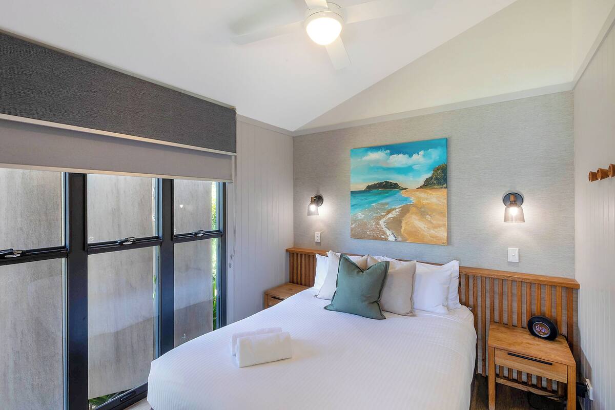 A bedroom with a large bed, two bedside tables, a painting of a beach, and a window with blinds.