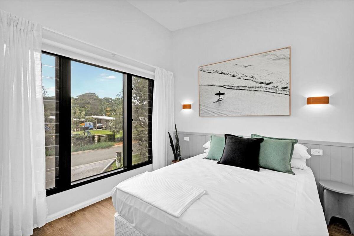 A bedroom with a large window, a bed with white bedding, and a painting of a snowy landscape.