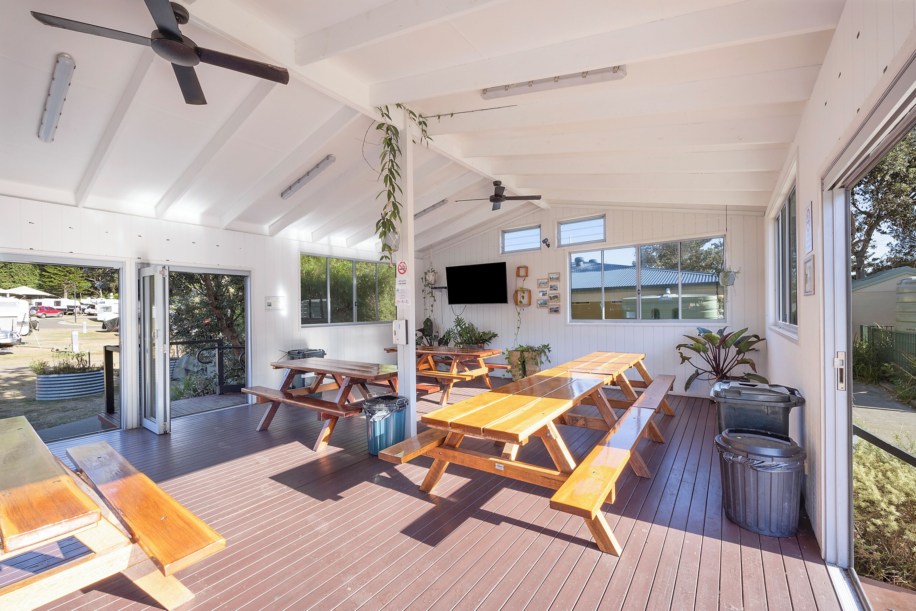 An outdoor patio with wooden picnic tables and benches, ceiling fans, and potted plants.