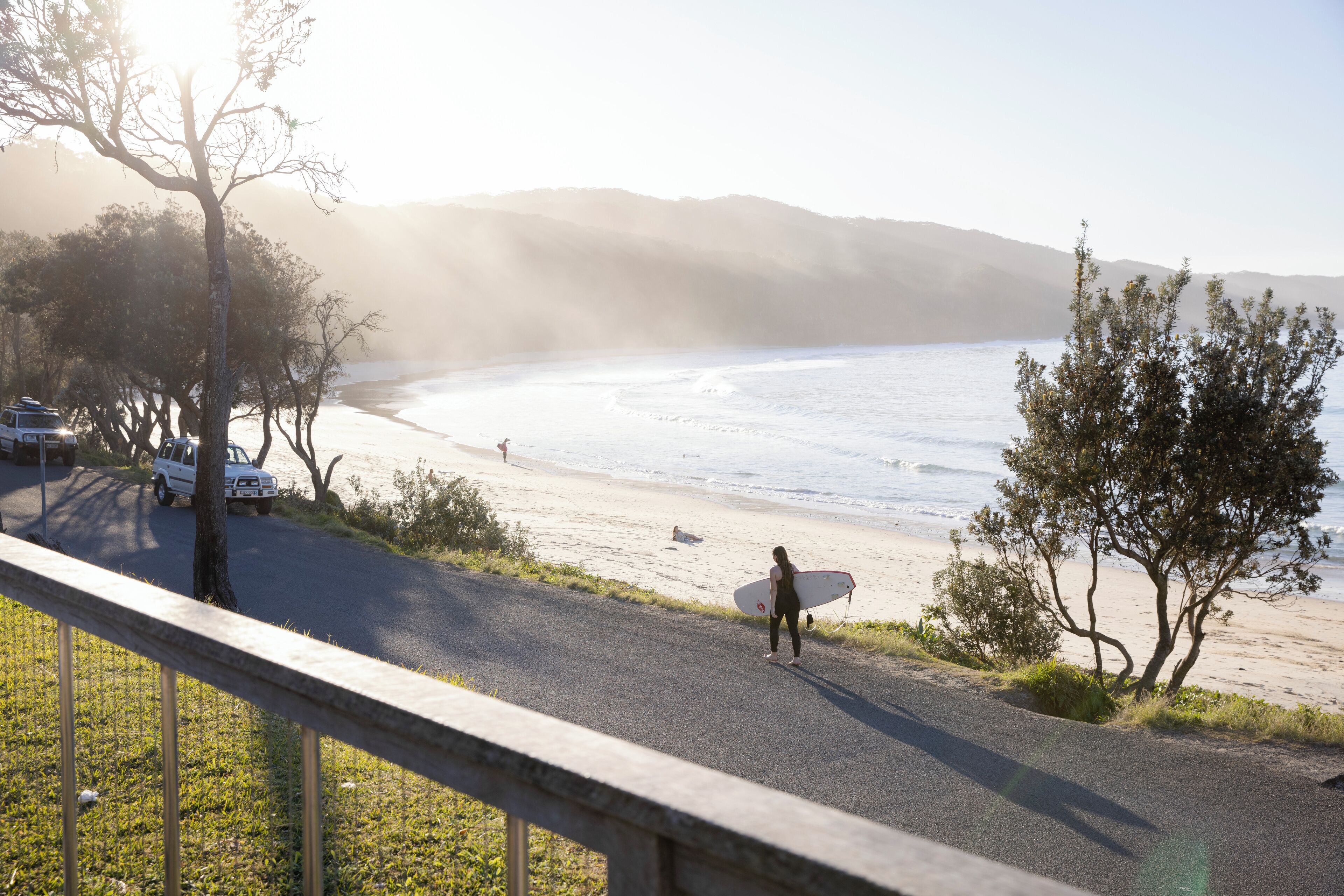 A coastal road with a person carrying a surfboard, a beach, and a mountainous backdrop.