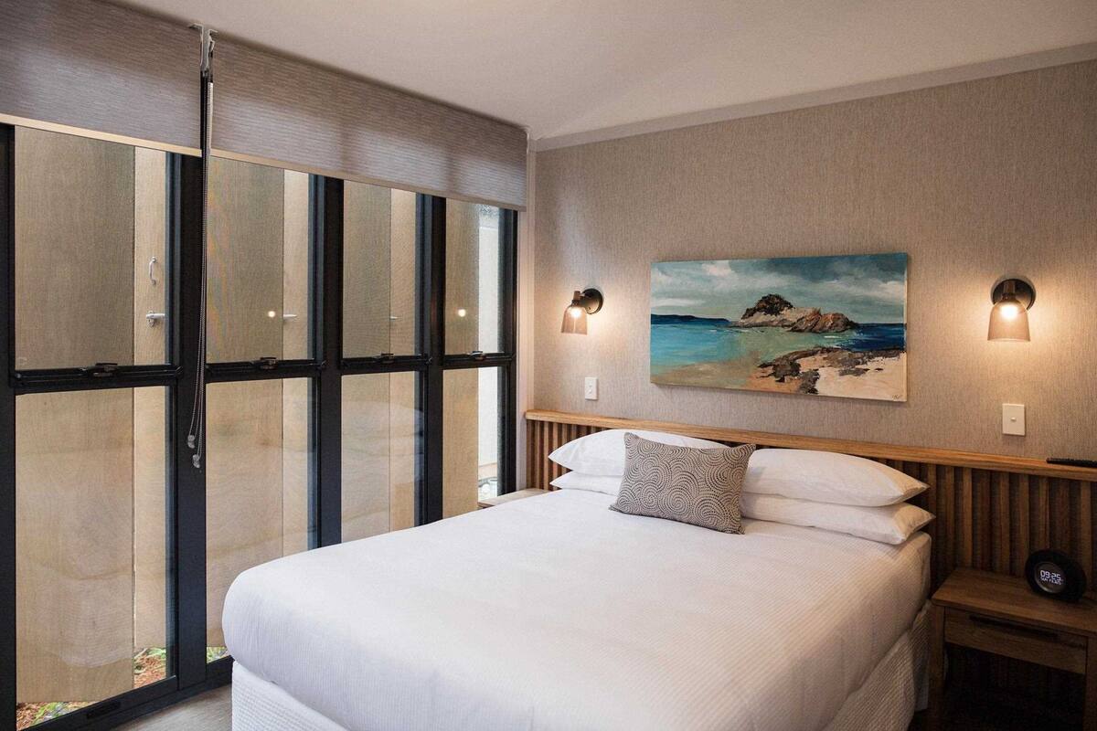A modern hotel room with a large bed, a painting of a coastal scene, and a sliding glass door leading to a balcony.