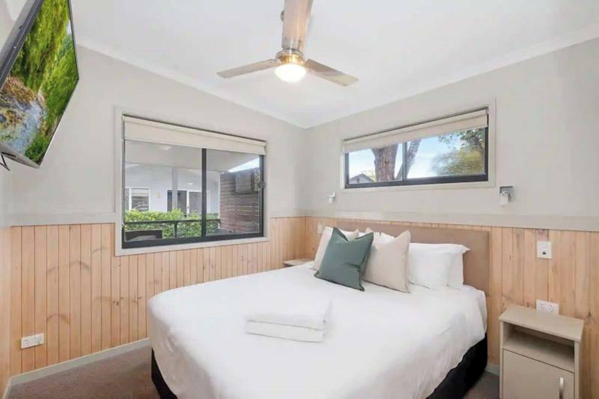 A bedroom with a bed, a ceiling fan, a window, and a TV mounted on the wall.