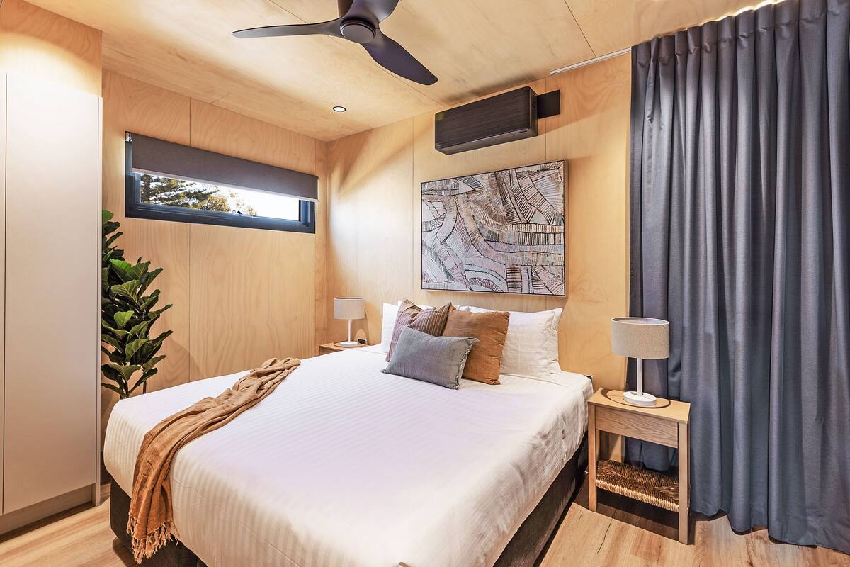 A modern bedroom with a large bed, a ceiling fan, a window with a view, a wall-mounted air conditioner, and a bedside table with a lamp.