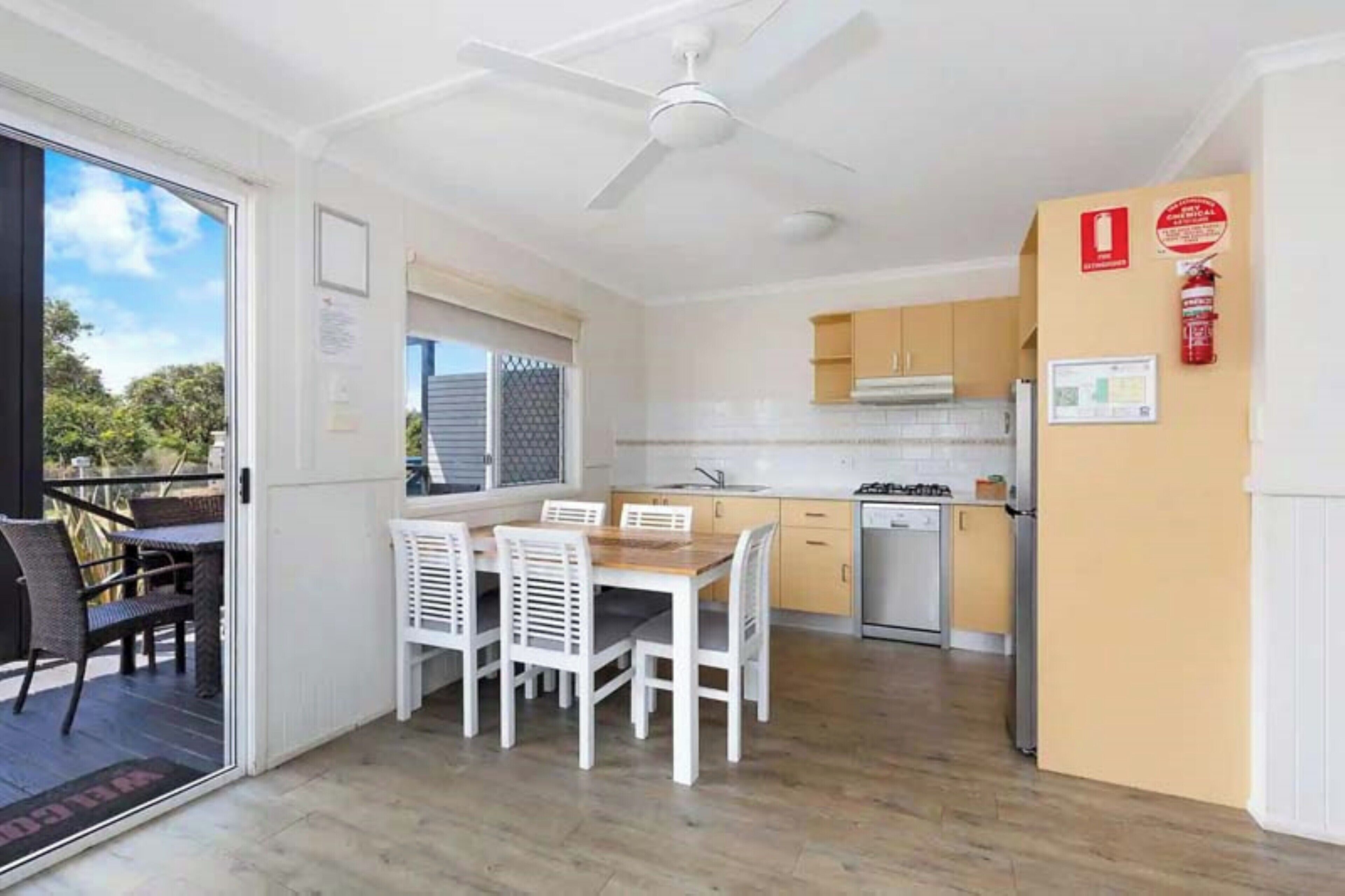 A compact kitchen with white cabinets, a dining table with chairs, a ceiling fan, and a fire extinguisher on the wall.