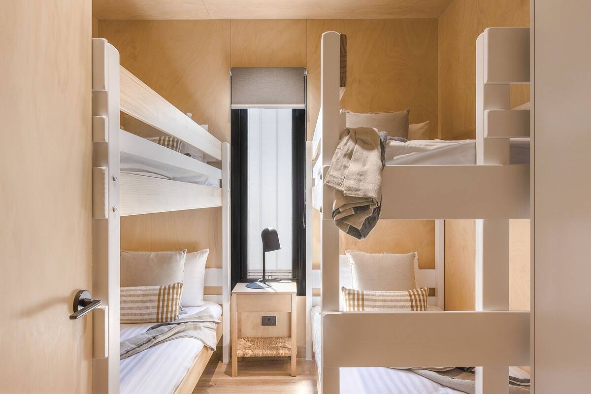 A compact, well-lit room with bunk beds, a small bedside table with a lamp, and a window providing natural light.