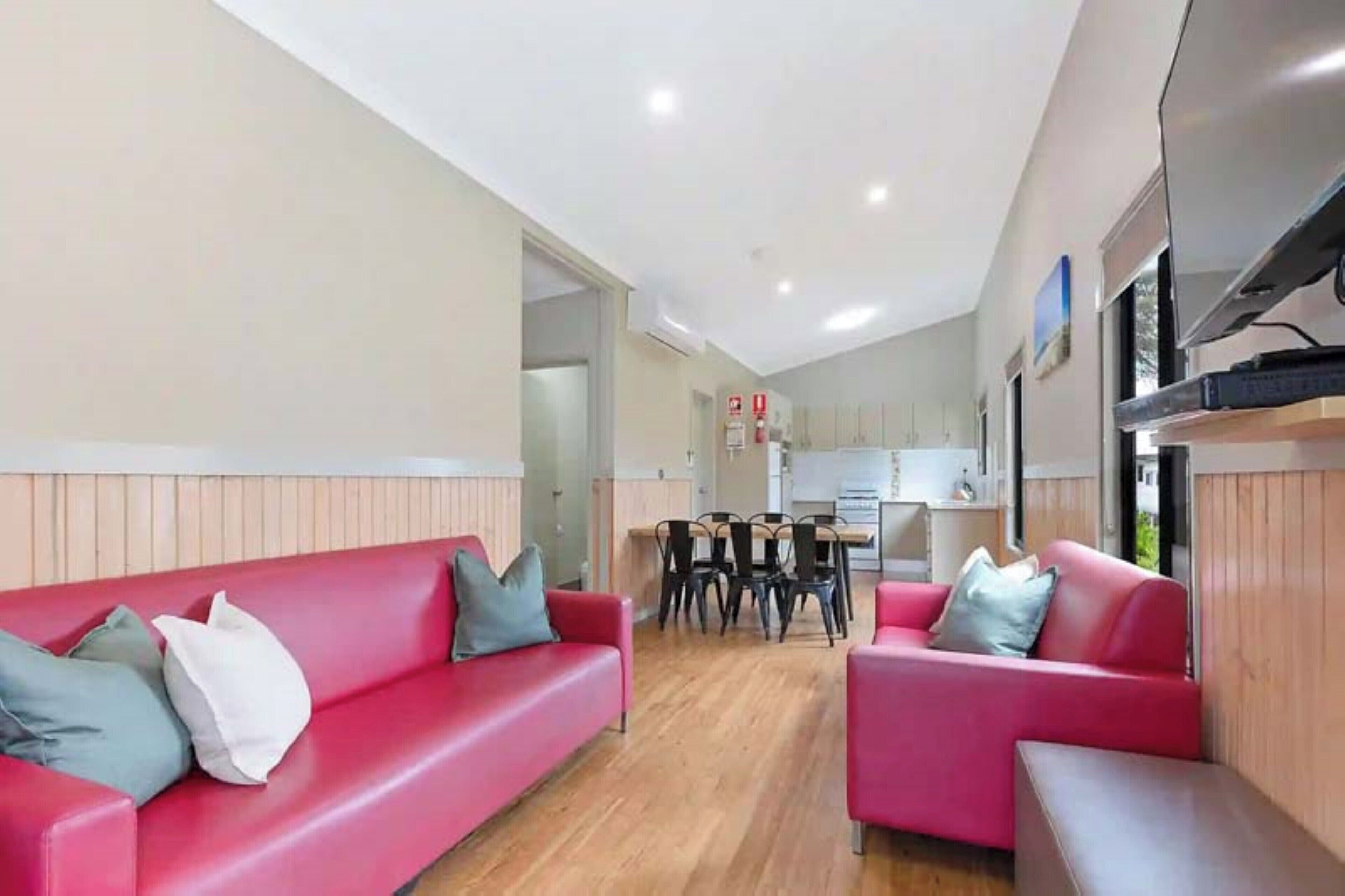 Un salon moderne avec deux canapés rouges, une table à manger en bois avec des chaises, et un coin cuisine à l’arrière-plan.
