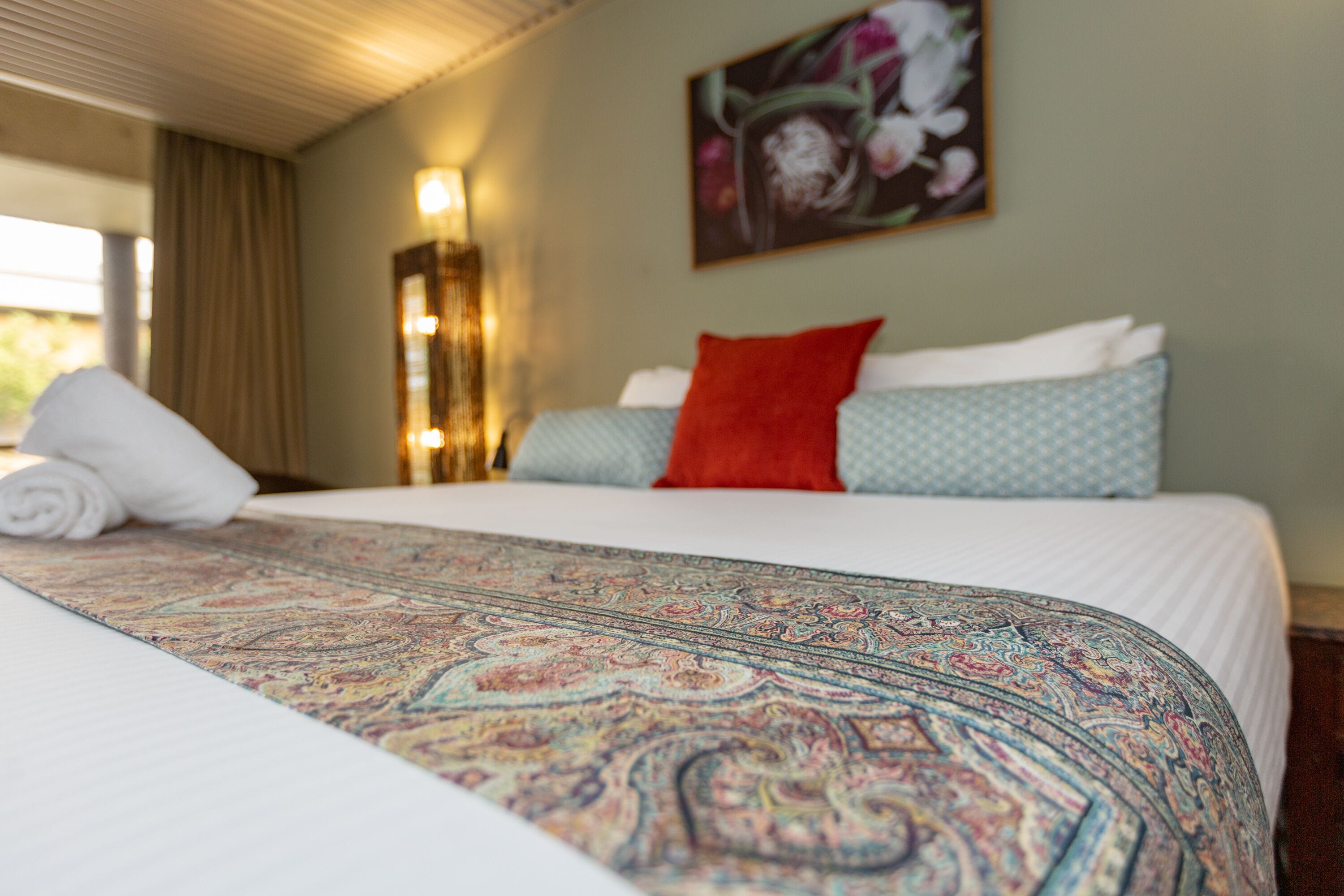 Una cama bien hecha con un edredón estampado, una almohada roja y una almohada azul sobre un colchón blanco.