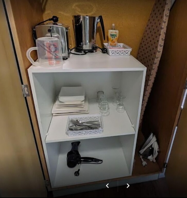 Una kitchenette compacta con cafetera, hervidor, refrigerador pequeño y un estante con utensilios y vasos.