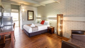 Una habitación de hotel moderna con suelo de madera, cama con ropa de cama blanca y almohada roja, escritorio con ordenador y pared de ladrillo.