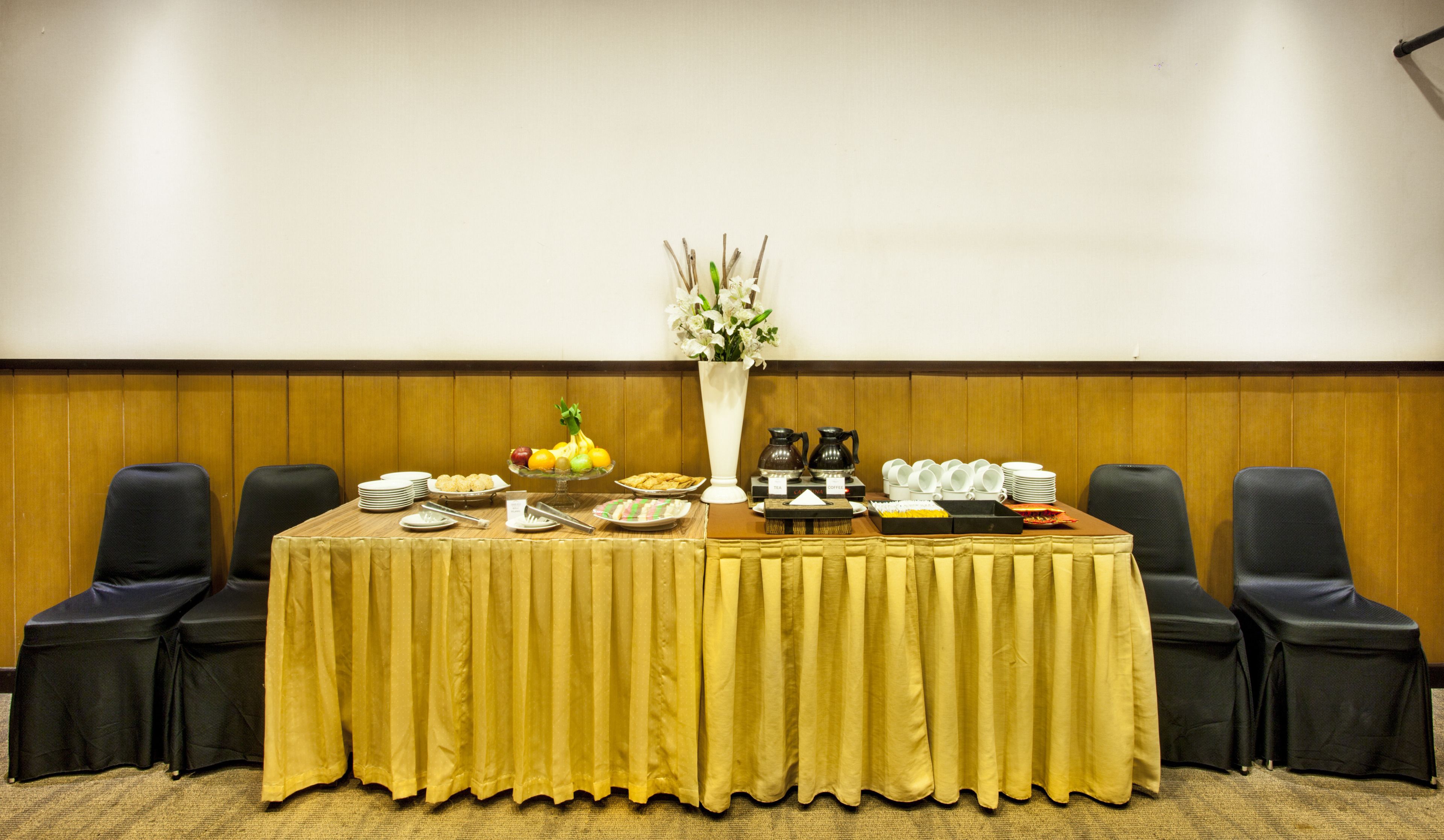 A conference table with a yellow tablecloth, surrounded by black chairs, with a white vase of flowers and various food and drink items on the table.