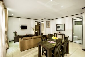 A modern living room with a sofa, dining table, and chairs. There is a TV mounted on the wall, a small cabinet, and a bathroom visible through a glass door.