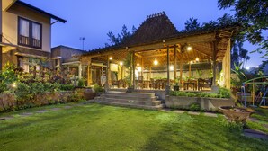 Un patio exterior bien iluminado con techo de paja, rodeado de abundante follaje y un césped cuidado.