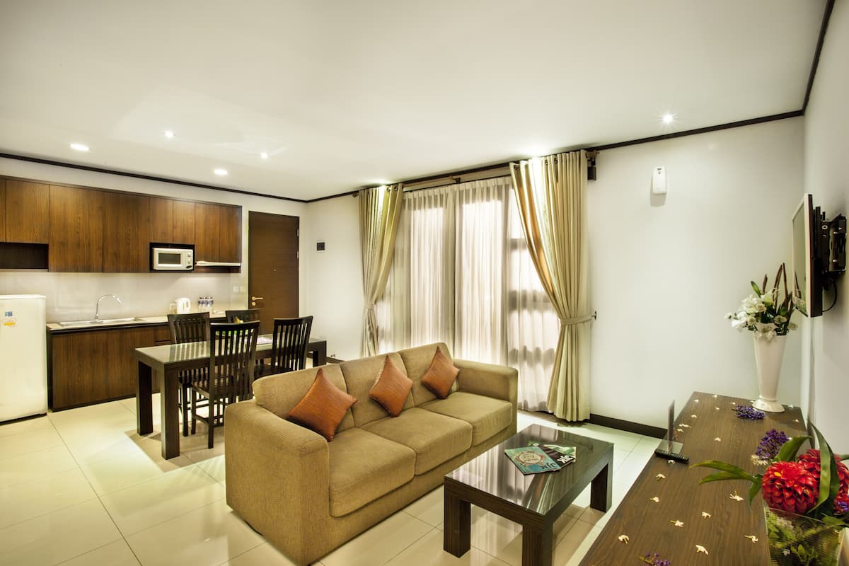 A modern living room with a sofa, dining table, and chairs. There is a kitchen area with a microwave and cabinets. The room has large windows with curtains.