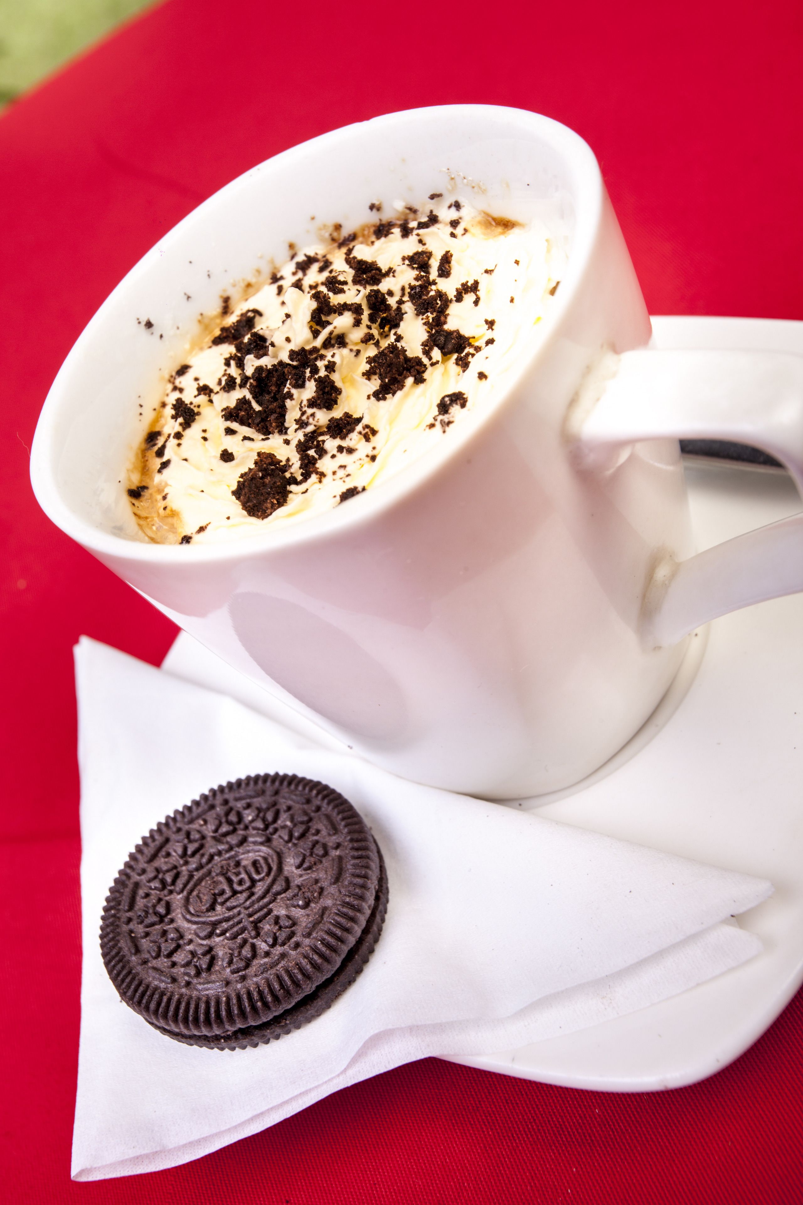 A white cup of coffee with whipped cream and chocolate crumbs, placed on a white napkin with an Oreo cookie beside it.