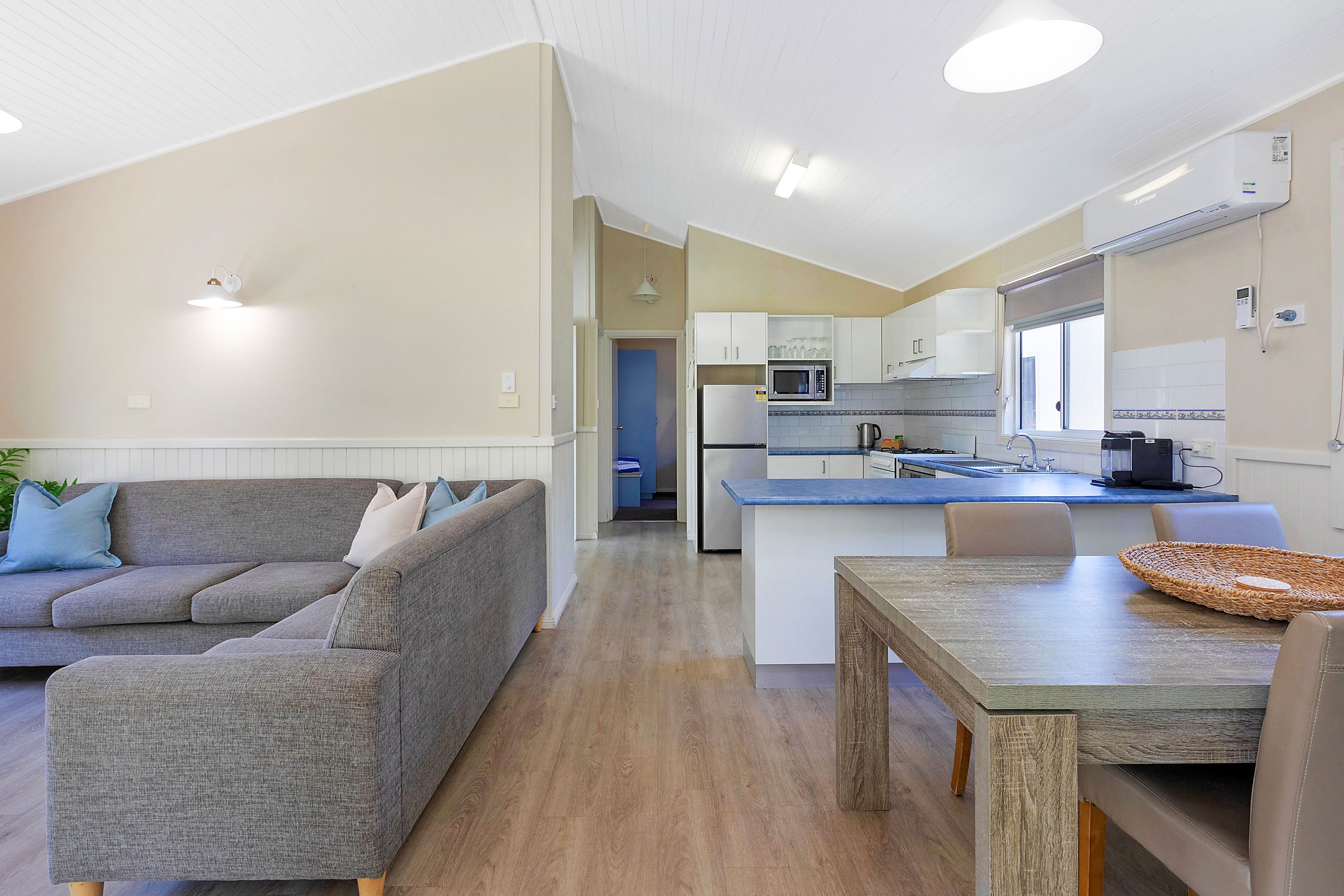 A modern living room with a grey sofa, a wooden dining table, and a kitchen area with white cabinets and a microwave.