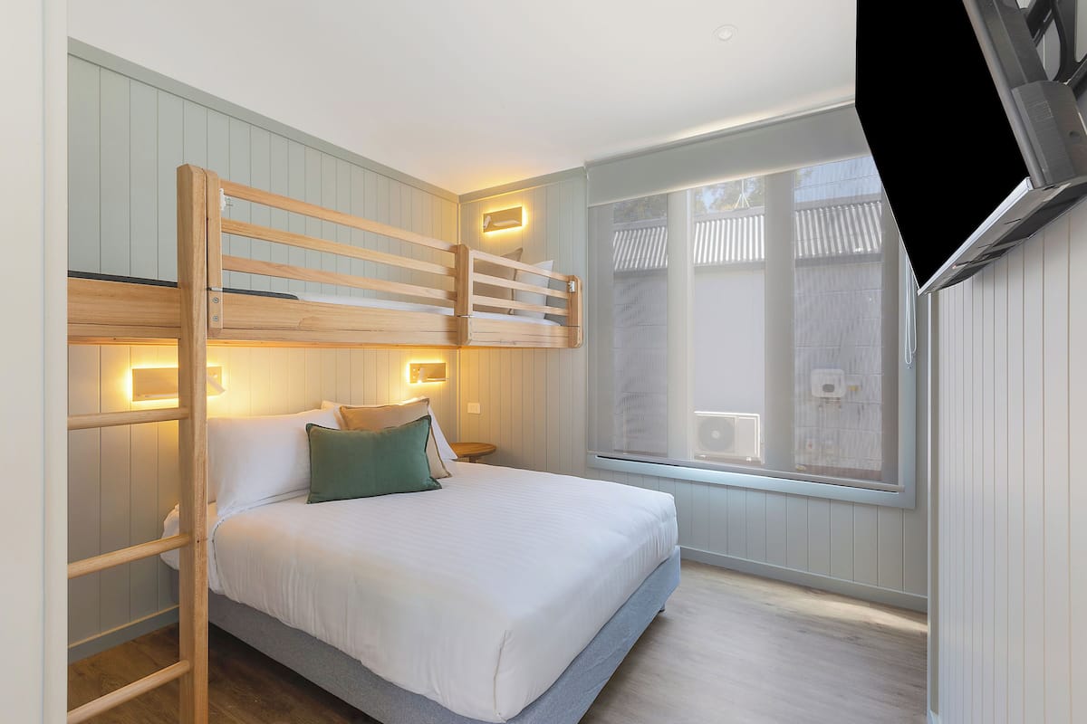 A bunk bed with a ladder, a bedside table, a wall-mounted light, and a window with blinds.