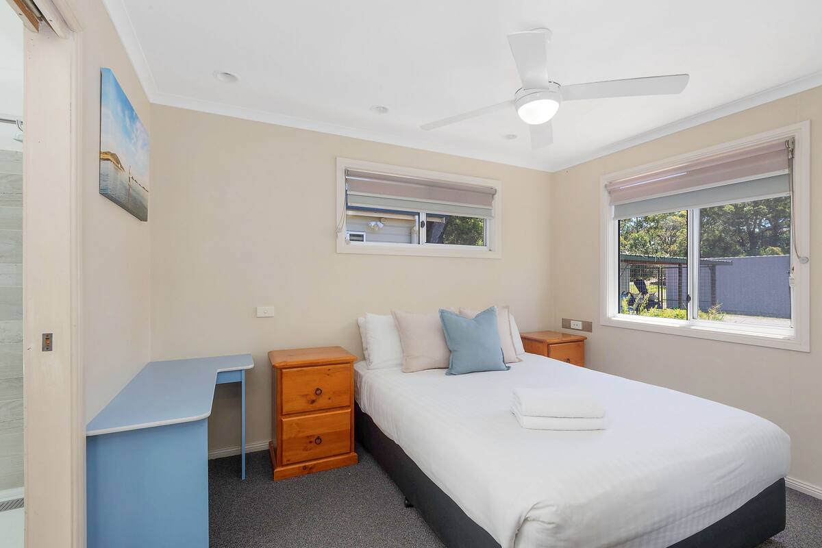 A bedroom with a bed, a desk, a dresser, a ceiling fan, and a window with blinds.