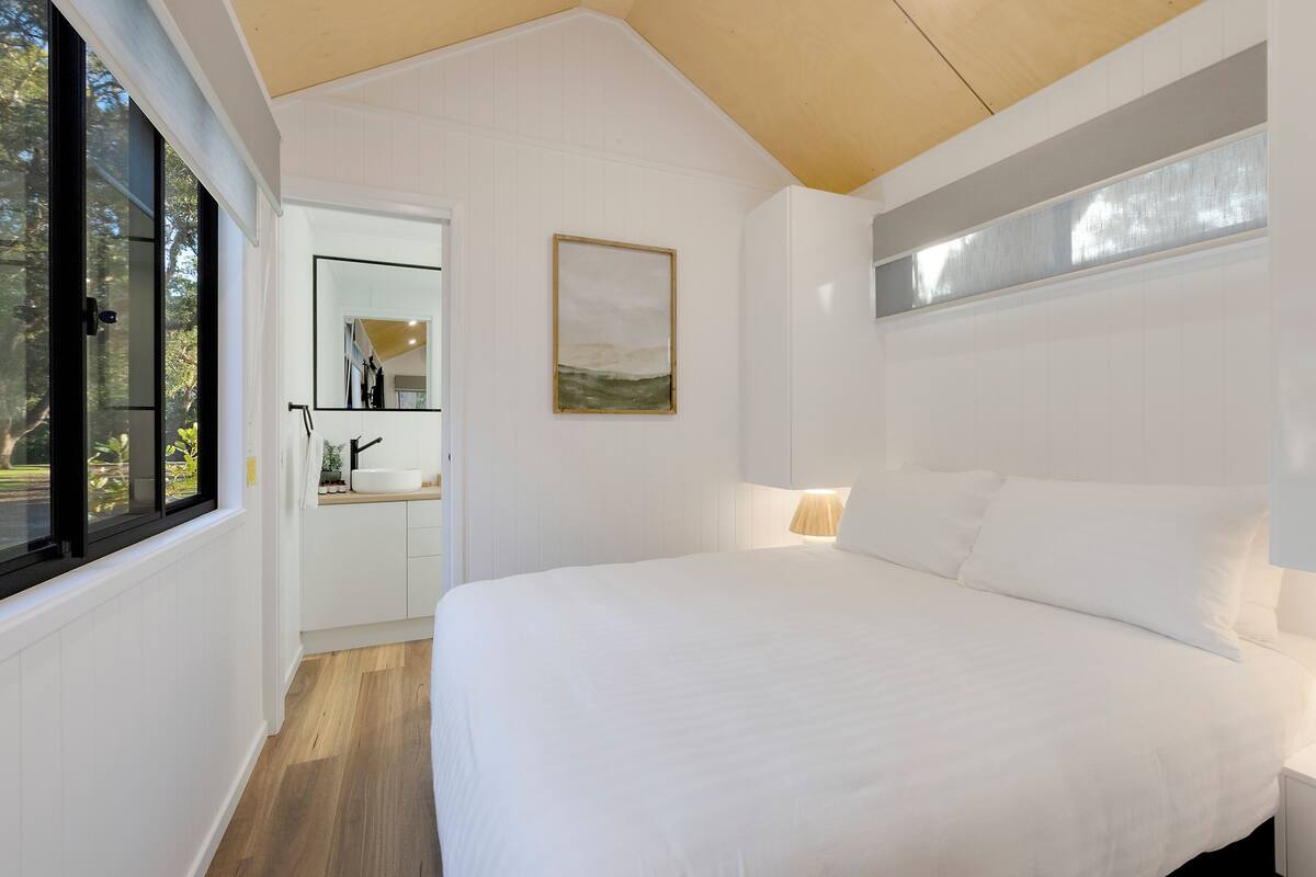 A bedroom with a large bed, a bedside table, a nightstand, a window, and a door leading to a bathroom.