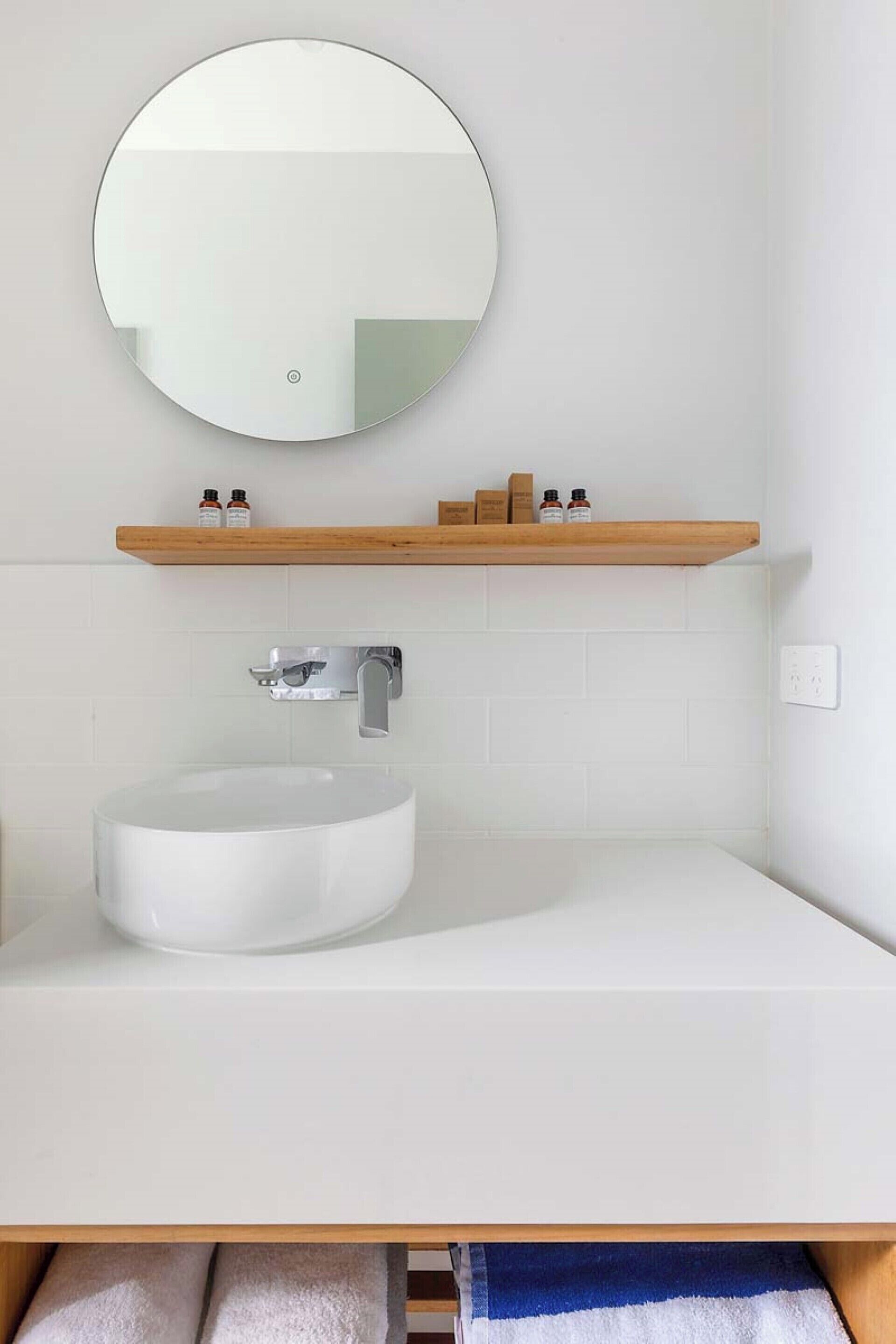 Un baño moderno con un espejo redondo, un lavabo blanco y una repisa de madera encima.