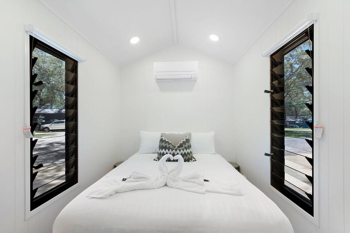 A hotel room with a bed, white walls, an air conditioning unit, and two windows with blinds.