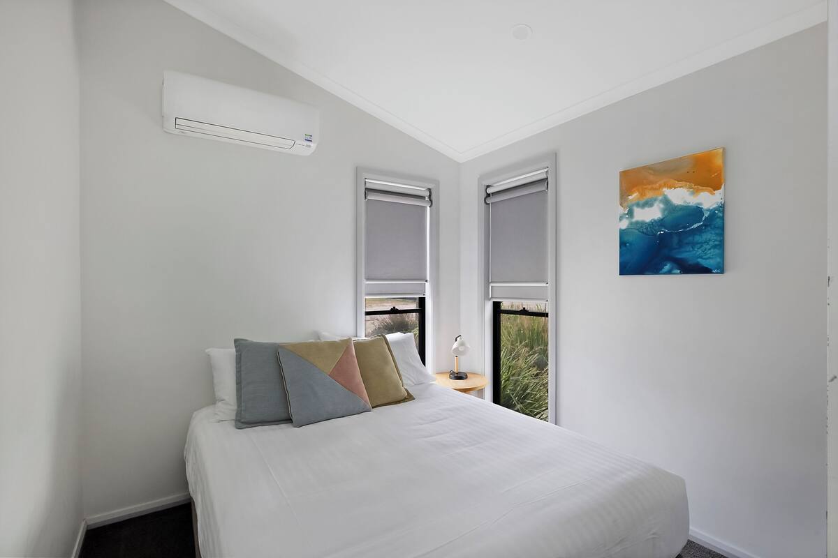 A bedroom with a bed, pillows, a nightstand, an air conditioner, and a window with a view.