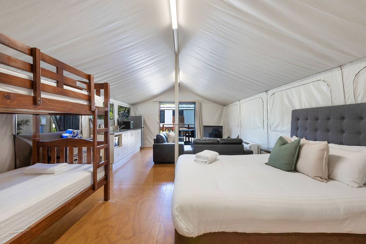 A cozy, well-lit room with a bunk bed, a large bed with pillows, a sofa, a TV, and a kitchenette area.