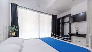 Habitación de hotel con cama, ventana con cortinas, televisor montado en la pared y una pequeña planta en una mesita de noche.