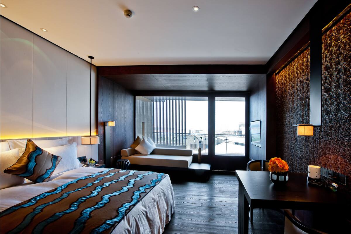 A modern hotel room with a large bed, a sofa, a dining table, and a view of the city through large windows.