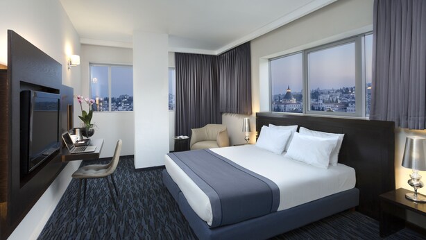 A modern hotel room with a large bed, a desk with a laptop, a chair, and a TV. - Golden Crown Old City Hotel (Nazareth)