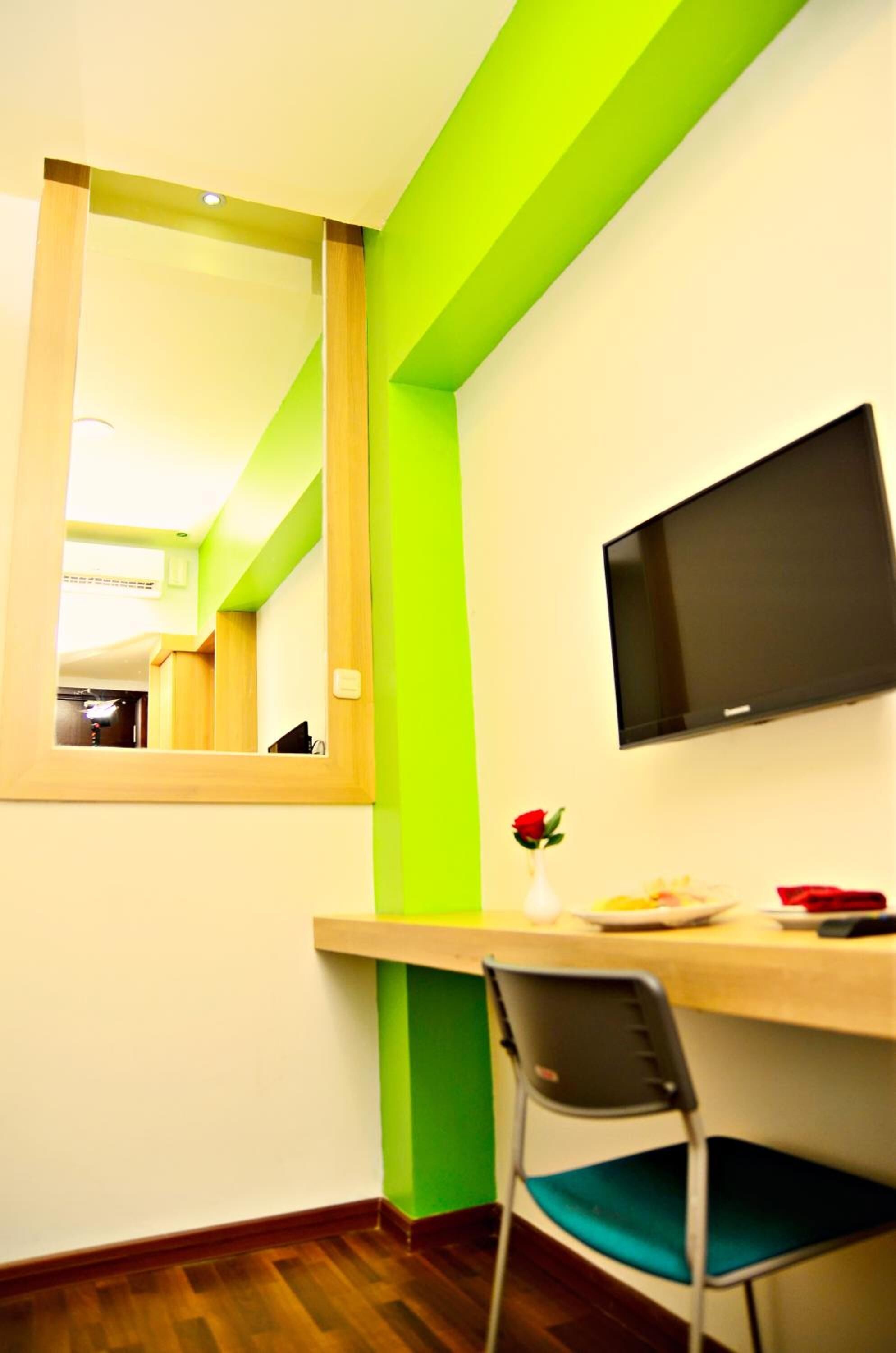 A room with a wooden desk, a chair, a television mounted on the wall, and a window with a mirror reflecting another room.
