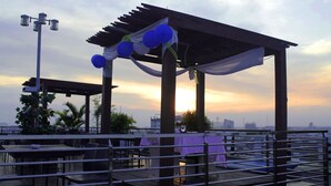 Un patio al aire libre con mesas y sillas, una pérgola adornada con globos azules y vistas al atardecer.