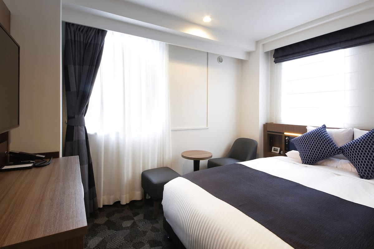 A hotel room with a bed, a desk, a chair, a small table, and a window with curtains.