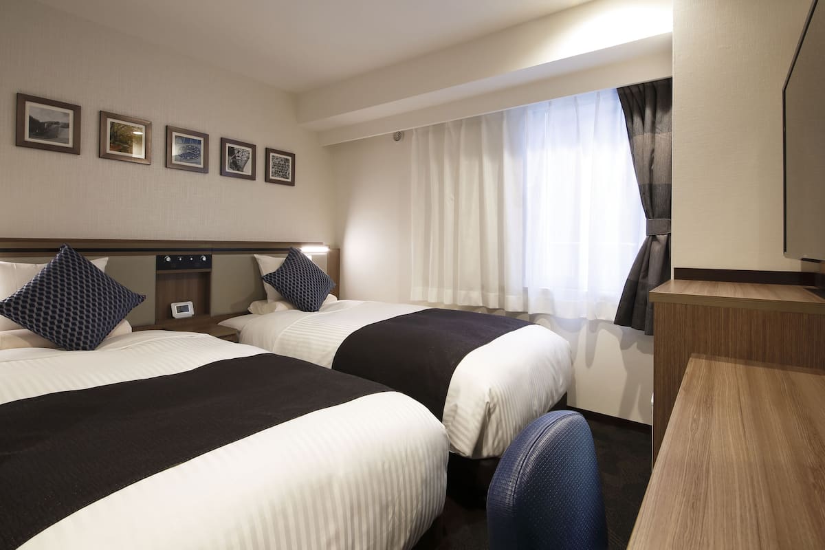 A hotel room with two beds, a desk, a TV, and a window with curtains.