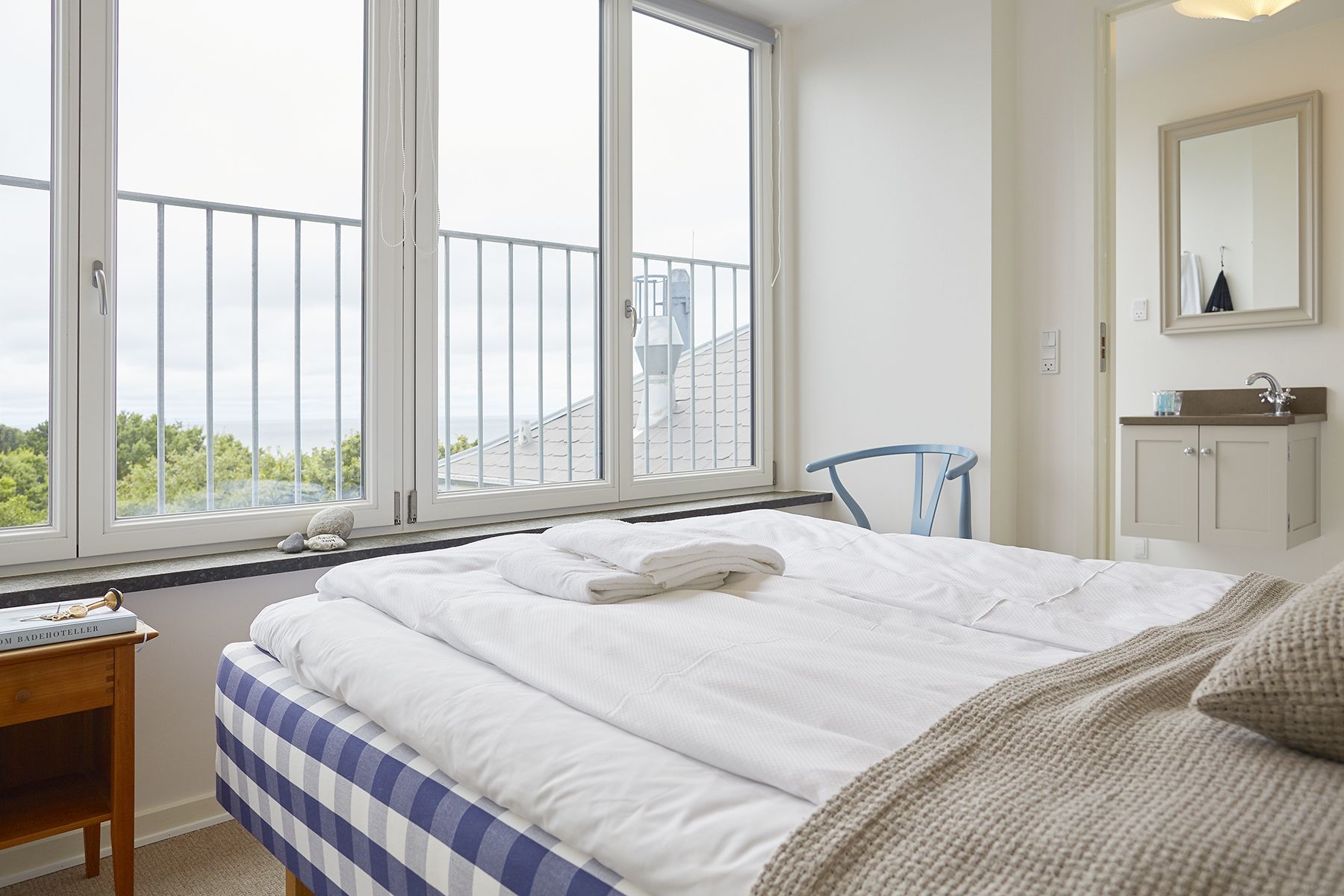 Una camera da letto con un'ampia finestra, un letto con biancheria da letto bianca, una sedia blu, un comodino con un libro e un bagno con uno specchio e un lavandino.