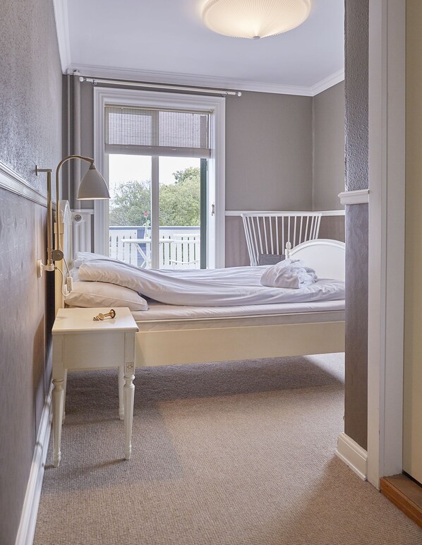 A bedroom with a single bed, a bedside table with a lamp, a window with blinds, and a view of a balcony. - Gilleleje Badehotel (Gilleleje)